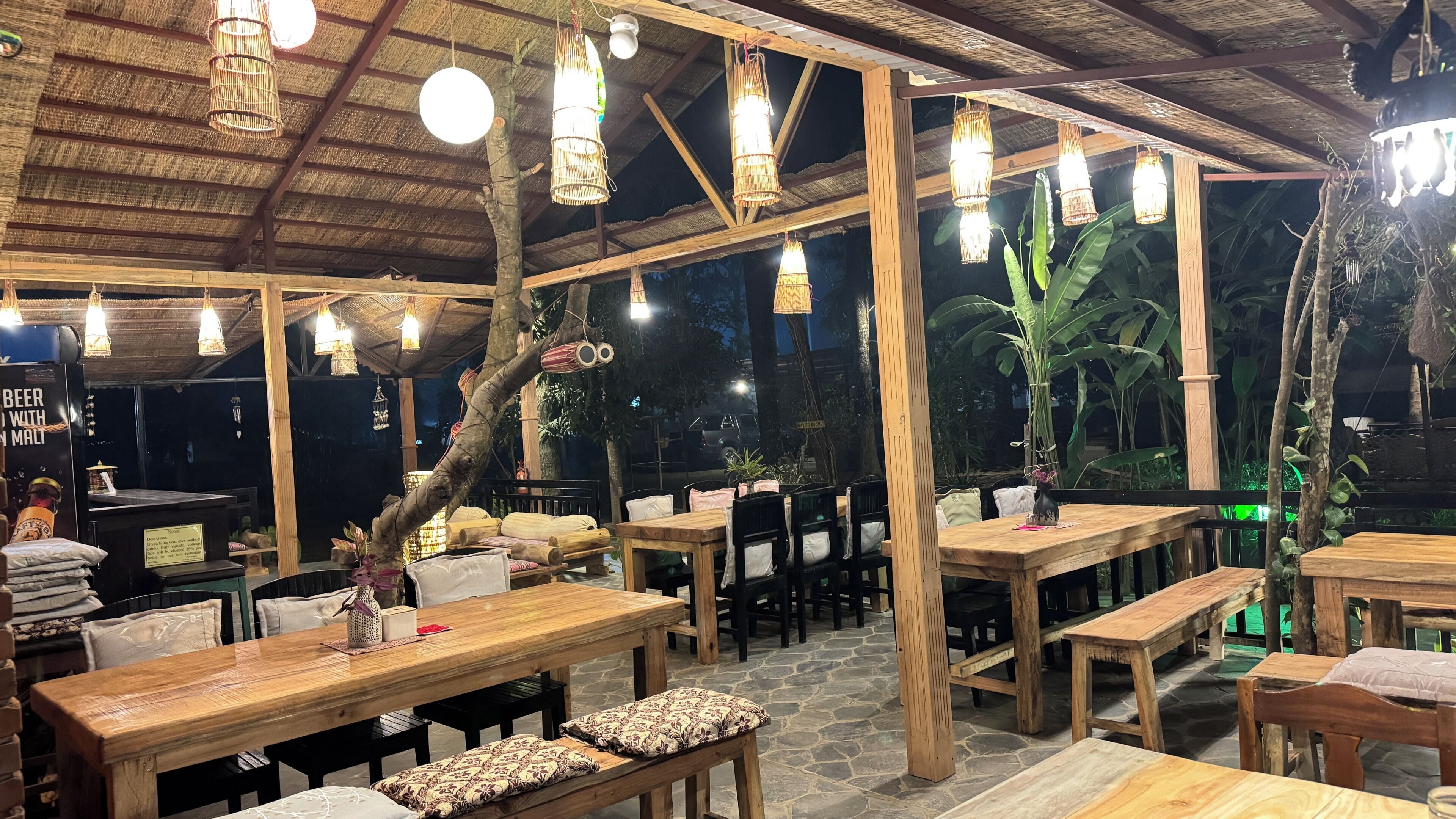 An outdoor restaurant with wooden tables and benches, hanging lights, and a thatched roof.