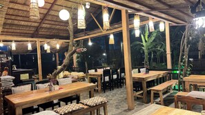 Un restaurante al aire libre con mesas y bancos de madera, luces colgantes y techo de paja.