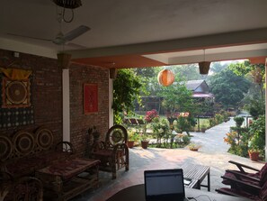 Una habitación con pared de ladrillo, ventilador de techo, una laptop sobre una mesa, mobiliario de mimbre y vista a un jardín con plantas en macetas y un edificio al fondo.