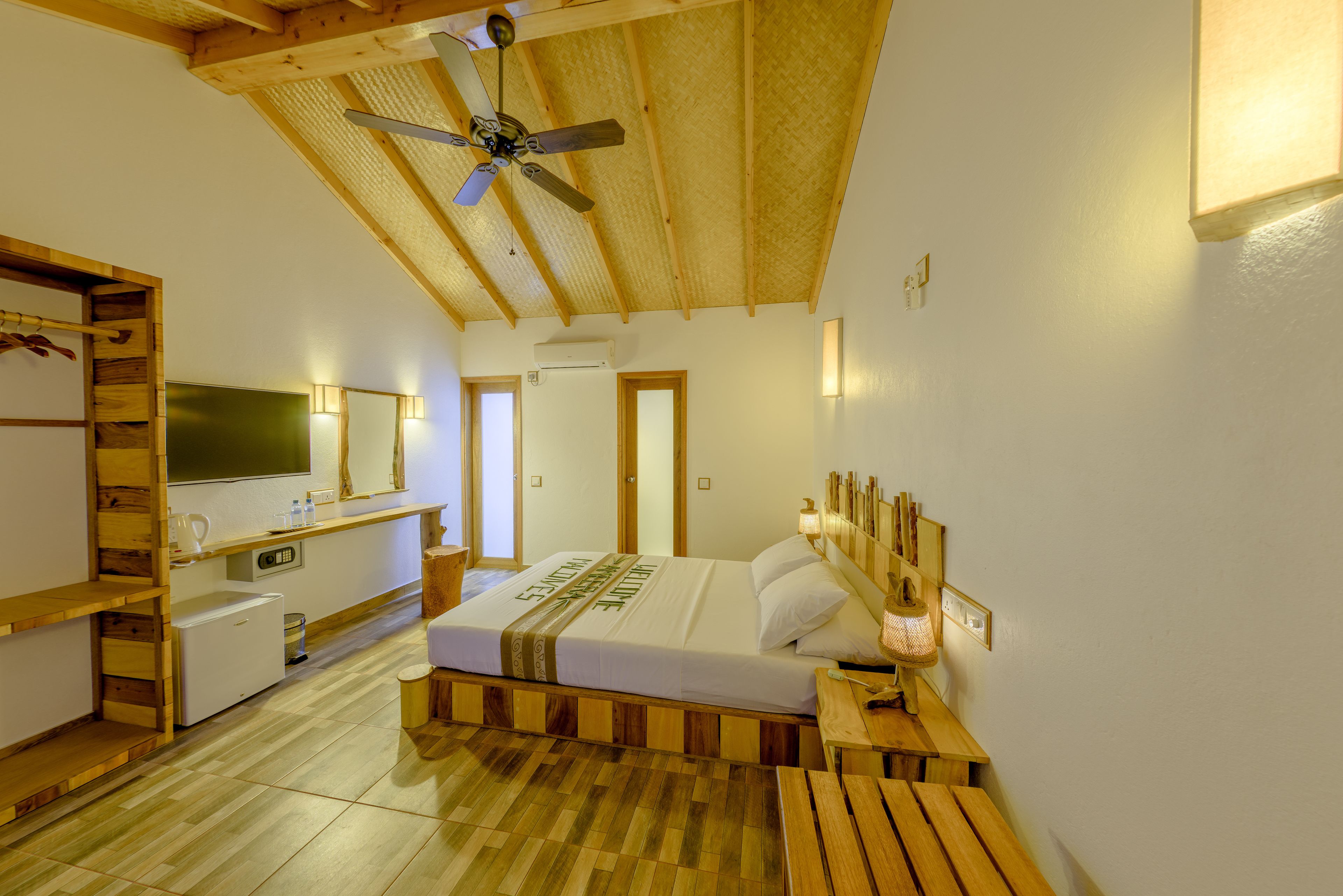A bedroom with a wooden ceiling, a bed with a headboard, a bedside table, a small desk, a television, and a wall-mounted shelf.