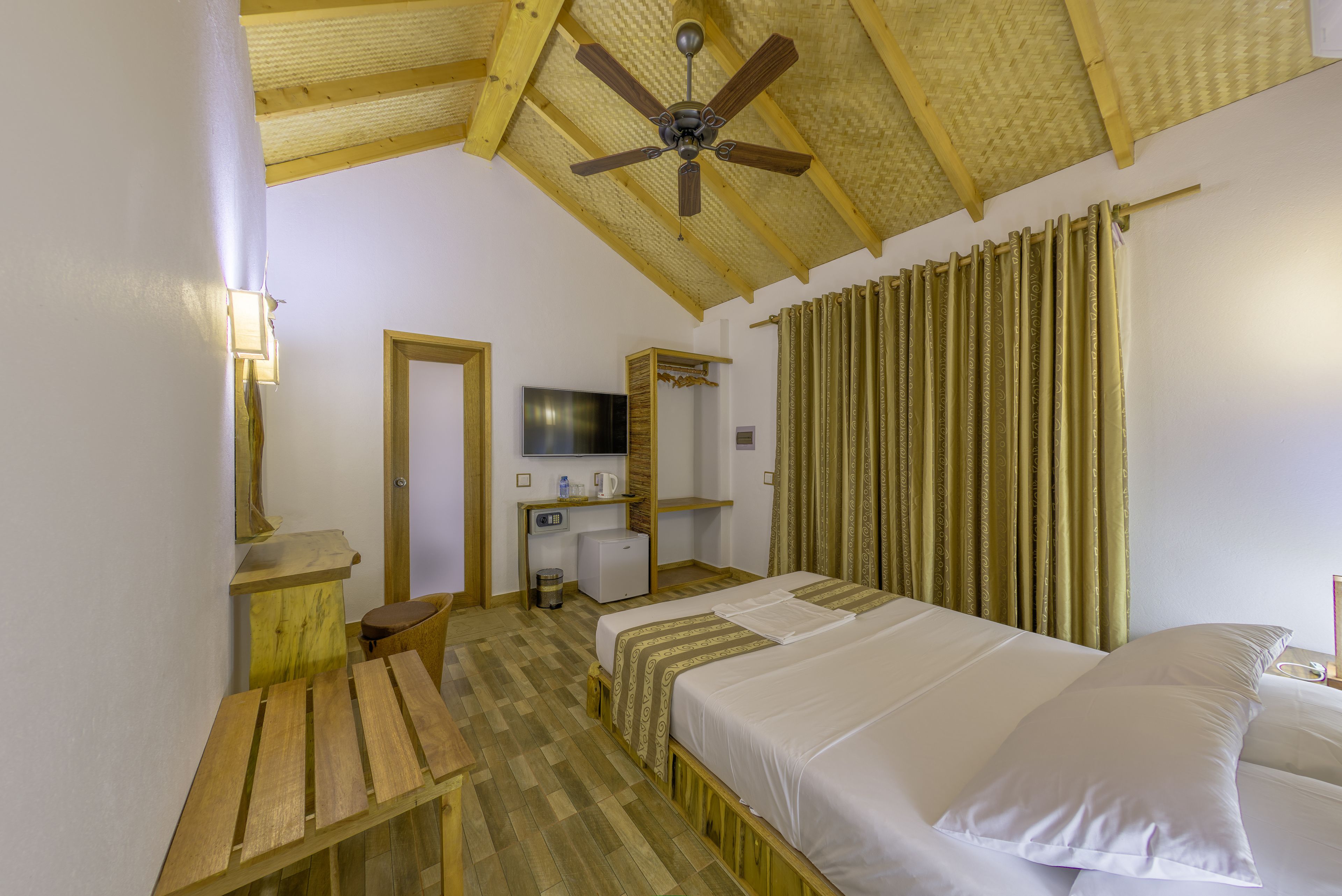 A hotel room with a wooden ceiling, a bed with white linens, a desk with a chair, a television, and a window with curtains.