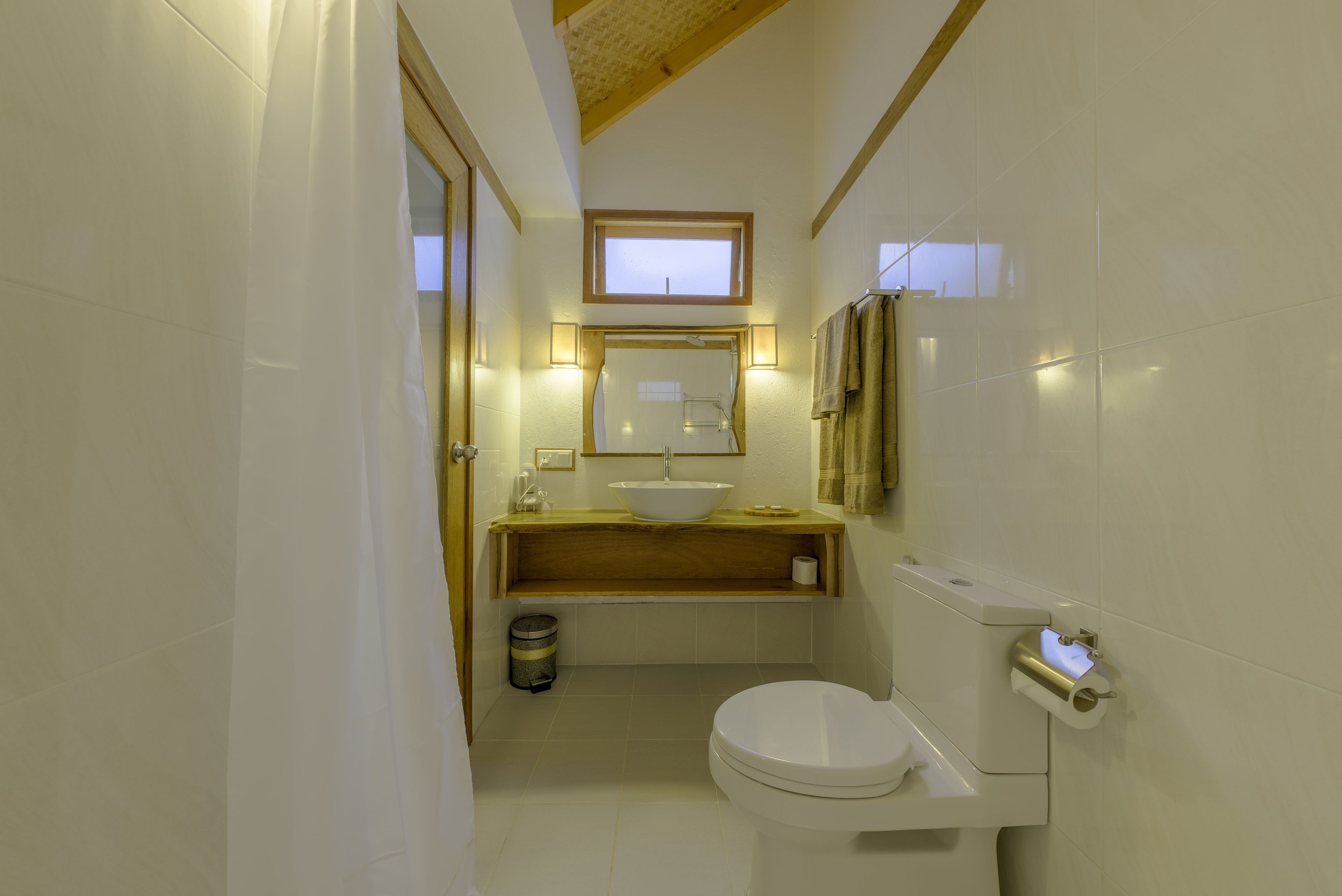 Une salle de bain avec des toilettes blanches, un meuble-lavabo en bois avec un évier, un miroir et une fenêtre située au-dessus du miroir.