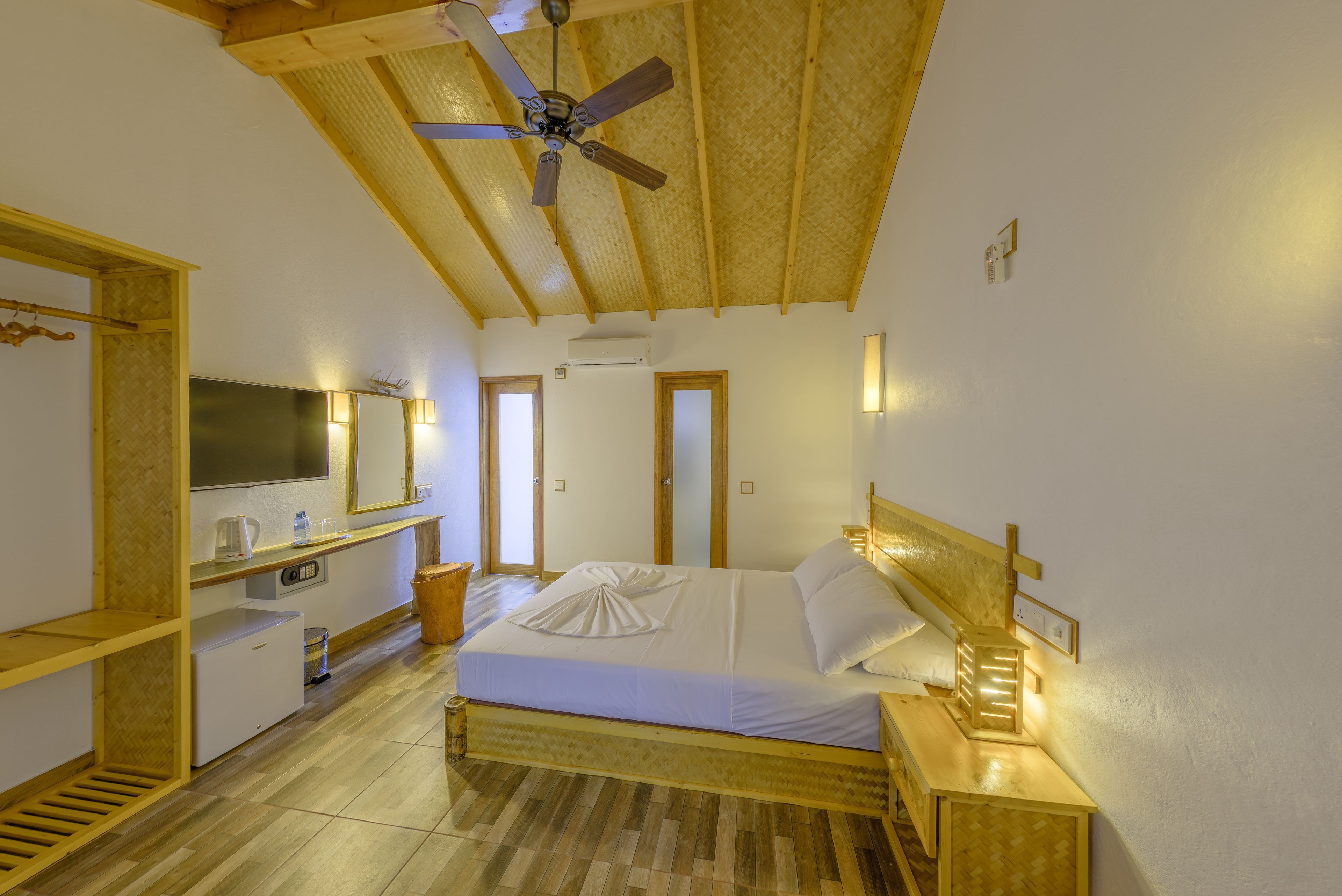 A bedroom with a bed, bedside tables, a ceiling fan, a TV, and a small kitchenette.