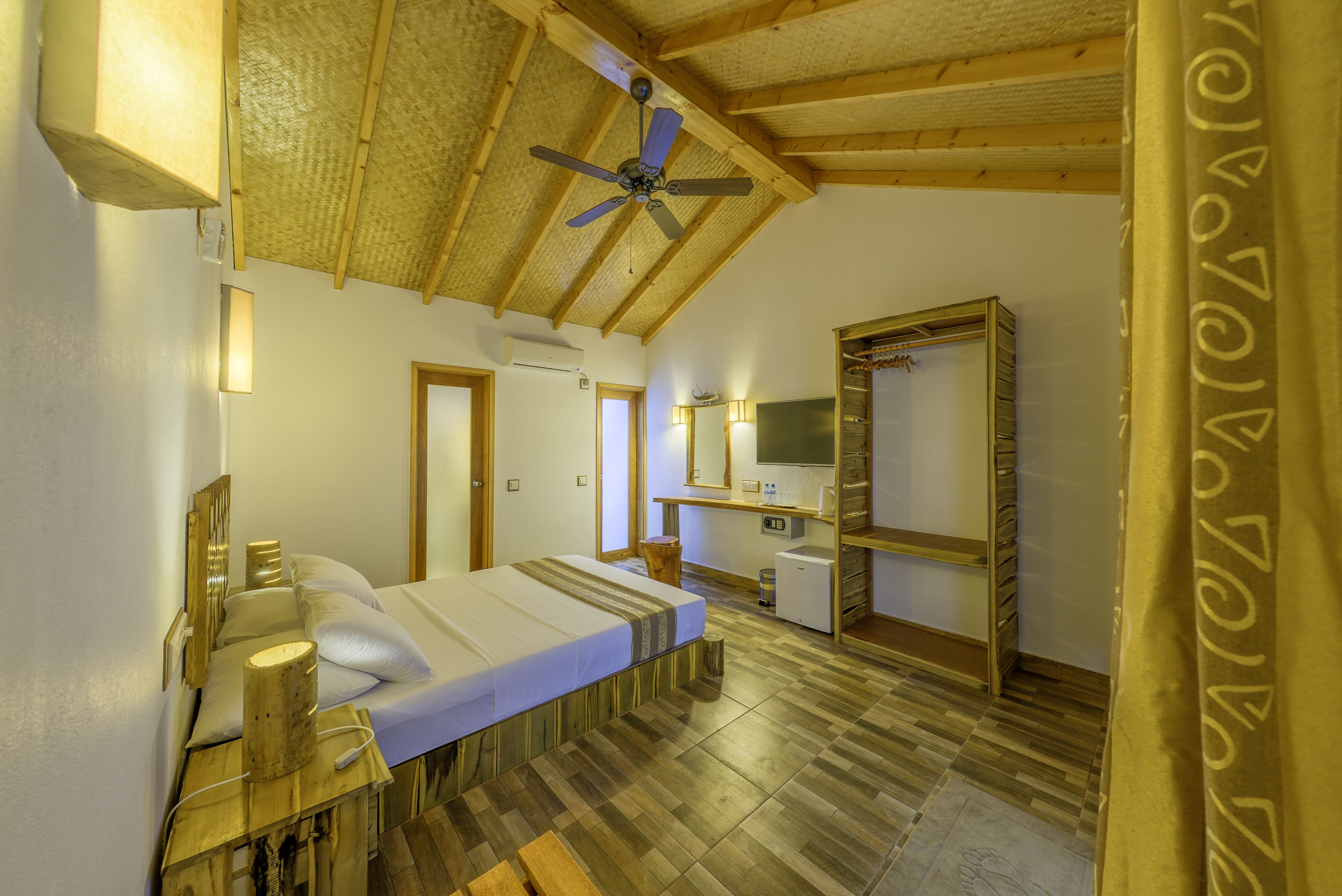 A room with a bed, wooden ceiling beams, a ceiling fan, a small kitchenette, and a mirror.