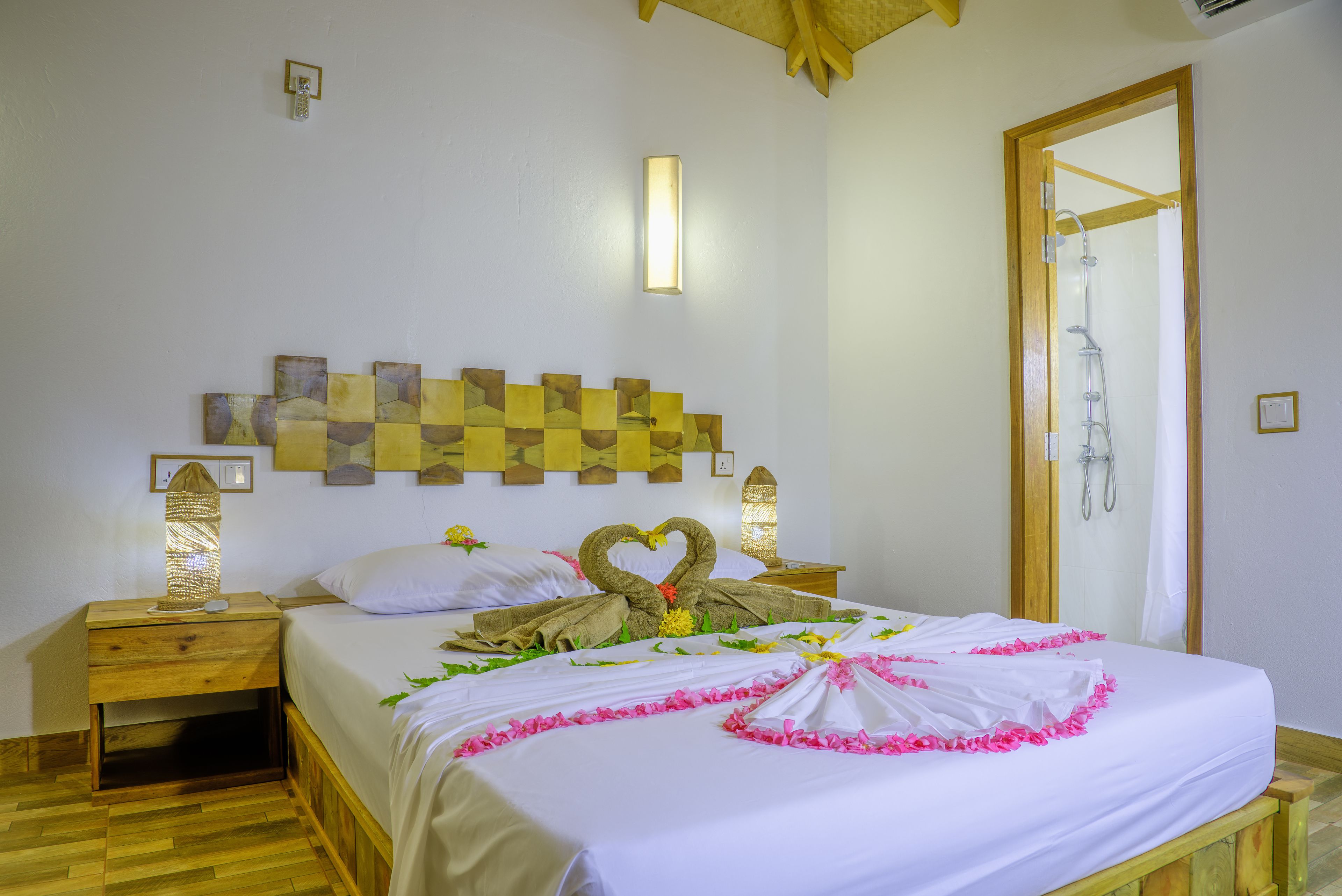 A neatly made bed with a heart-shaped towel arrangement, a wooden headboard with a geometric pattern, and a wooden nightstand with a lamp.