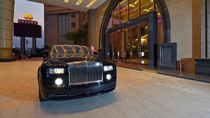 A luxury car parked in front of a hotel entrance with a large illuminated sign and a grand arched doorway. - Huilihua Hotel (Dongguan)
