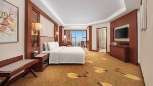 A hotel room with a bed, a desk, a chair, a TV, and a view of the city. - Huilihua Hotel (Dongguan)