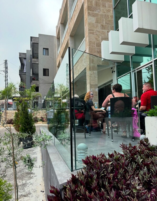 An outdoor seating area with glass railings, potted plants, and a view of a modern building with multiple balconies. - Byblostar Hotel (Byblos)