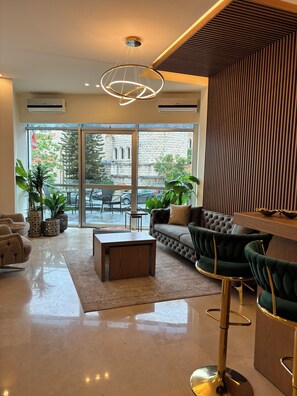 A modern living room with a sofa, chairs, a coffee table, and a large window with a view of a building and trees. - Byblostar Hotel (Byblos)