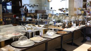 A buffet setup with various food items and serving utensils, a bar area with a person behind the counter, and hanging decorative lights. - Delora Hotel and Suites (Chtaura)