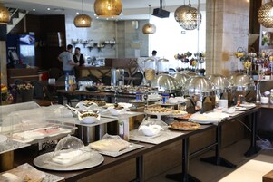 Disposição de buffet com diversos alimentos e utensílios, área de bar com uma pessoa atrás do balcão e luminárias decorativas penduradas.