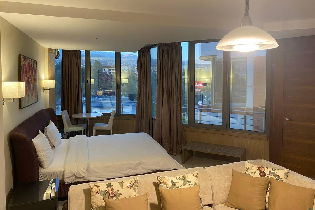 Une chambre d’hôtel équipée d’un lit, d’un bureau, d’une chaise, d’un canapé et de grandes fenêtres donnant sur la ville.