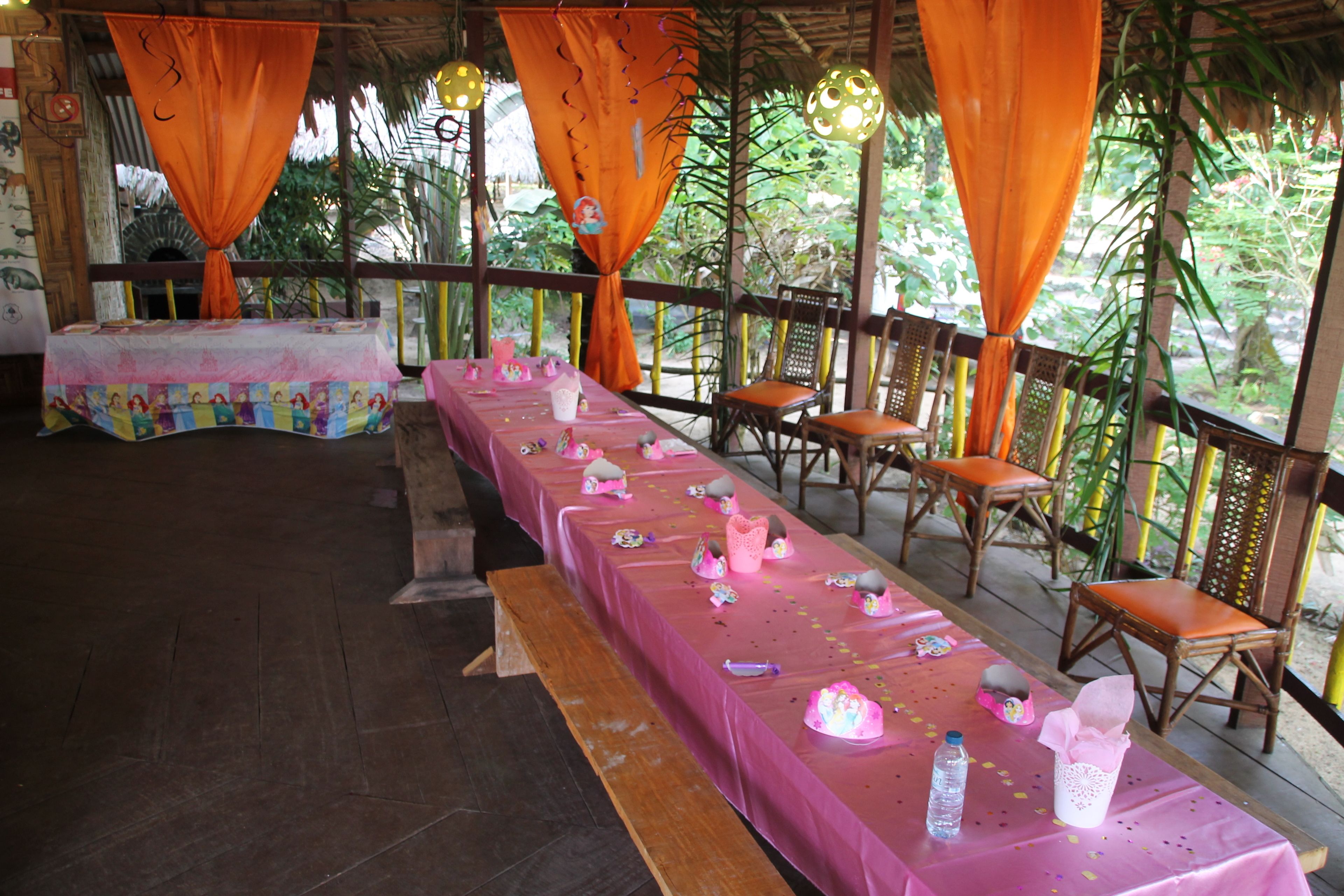 Une longue table recouverte de nappes roses, avec des rideaux orange, entourée de chaises en bois.