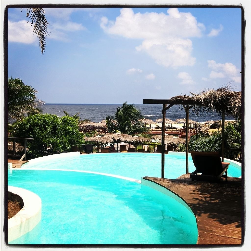 A pool area with a view of the ocean, surrounded by palm trees and thatched umbrellas.