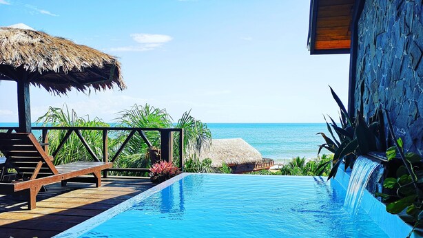 A pool with a view of the ocean, wooden lounge chairs, and a thatched roof structure. - Libassa Ecolodge (Marshall)