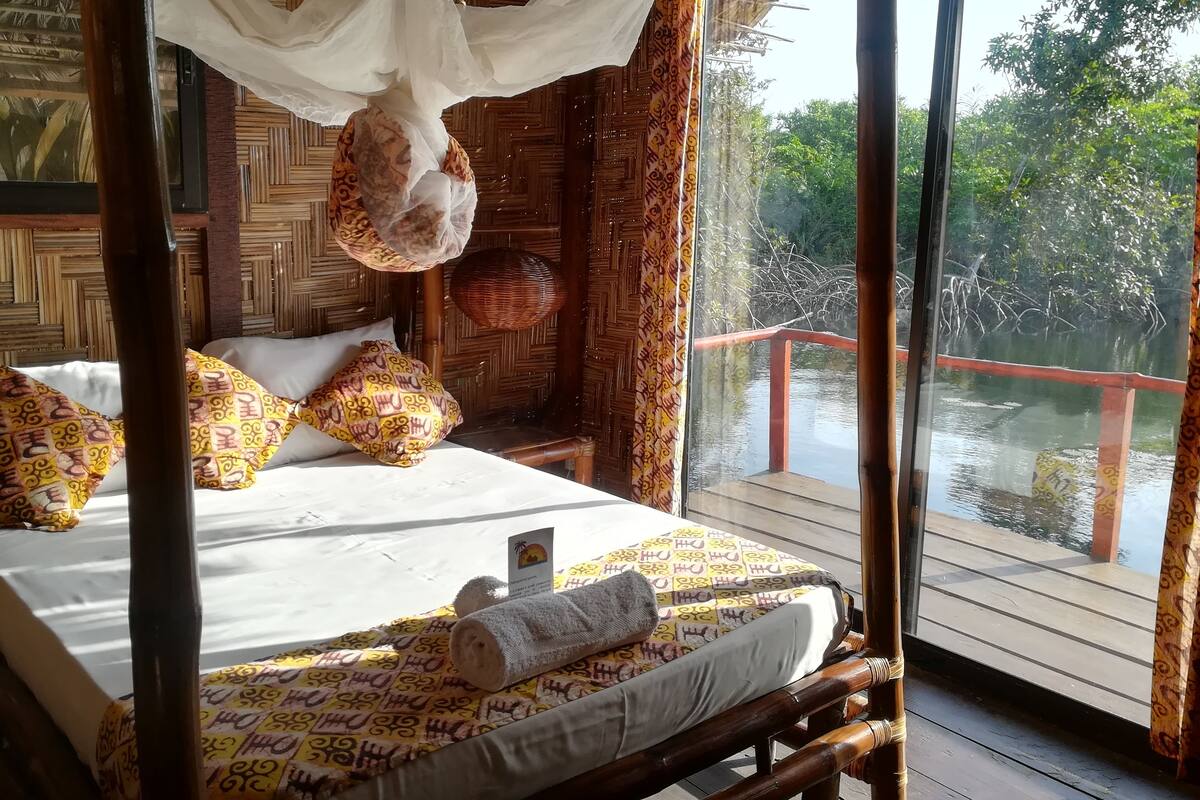 A room with a bed, pillows, a canopy, and a view of a water body.