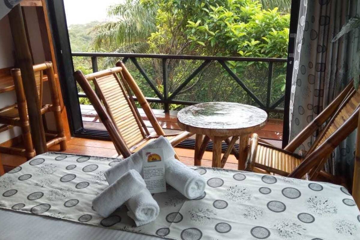 A balcony with wooden chairs and a small round table, a bed with a patterned bedspread, and a view of lush greenery.