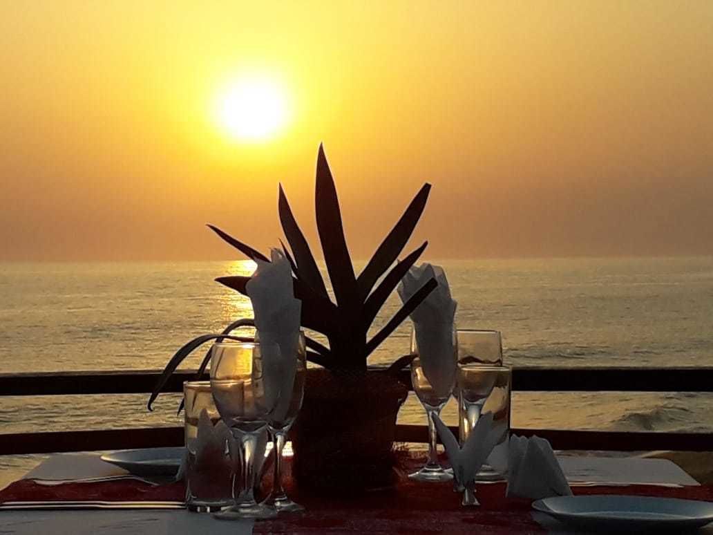 Una mesa para dos con planta en maceta, vasos y una vela, con vista a la puesta de sol sobre el océano.