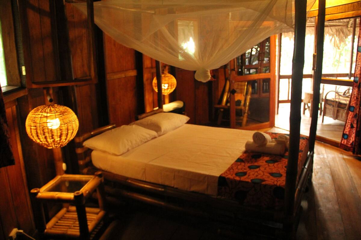 A wooden room with a bed, a chair, a hanging lamp, and a window with a view.