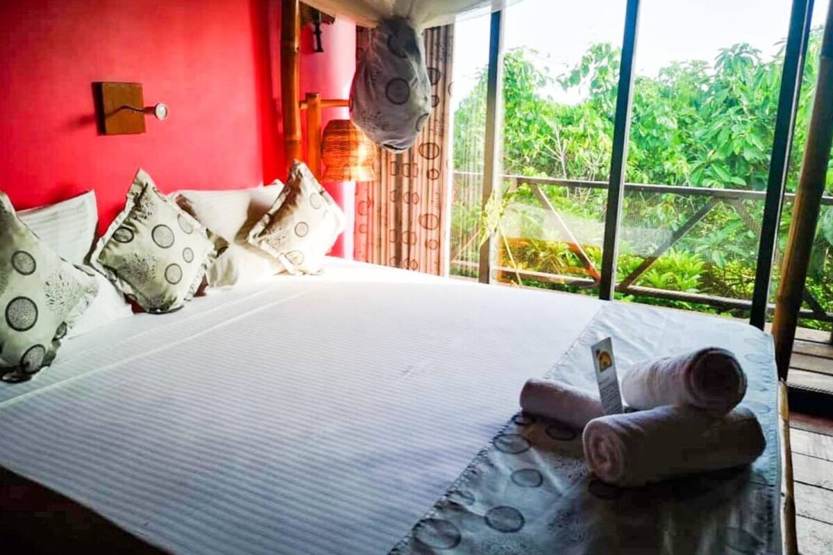 A bedroom with a large bed, pillows, a rolled towel, and a window with a view of greenery.