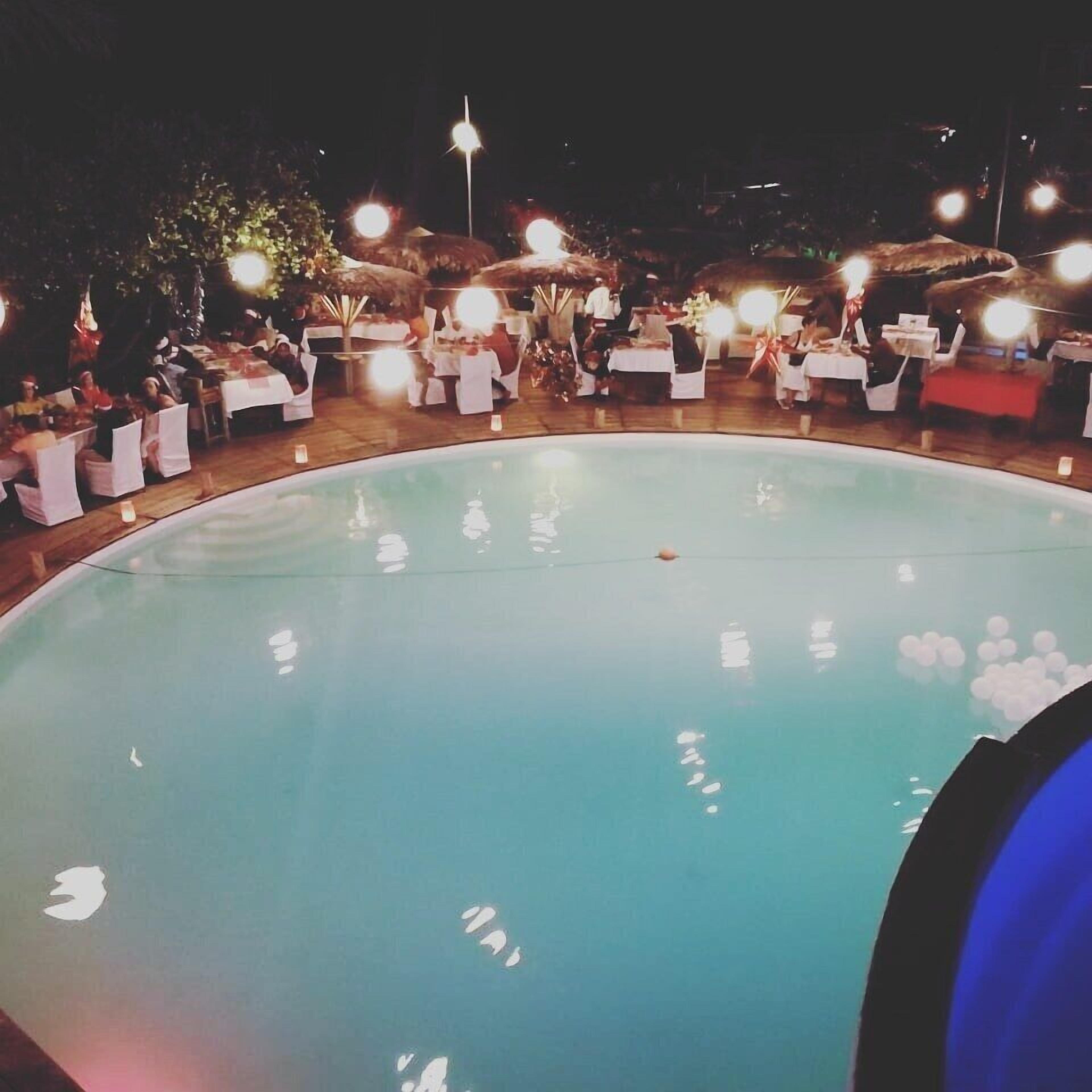 Un área junto a la piscina con mesas y sillas, rodeada de sombrillas y luces.