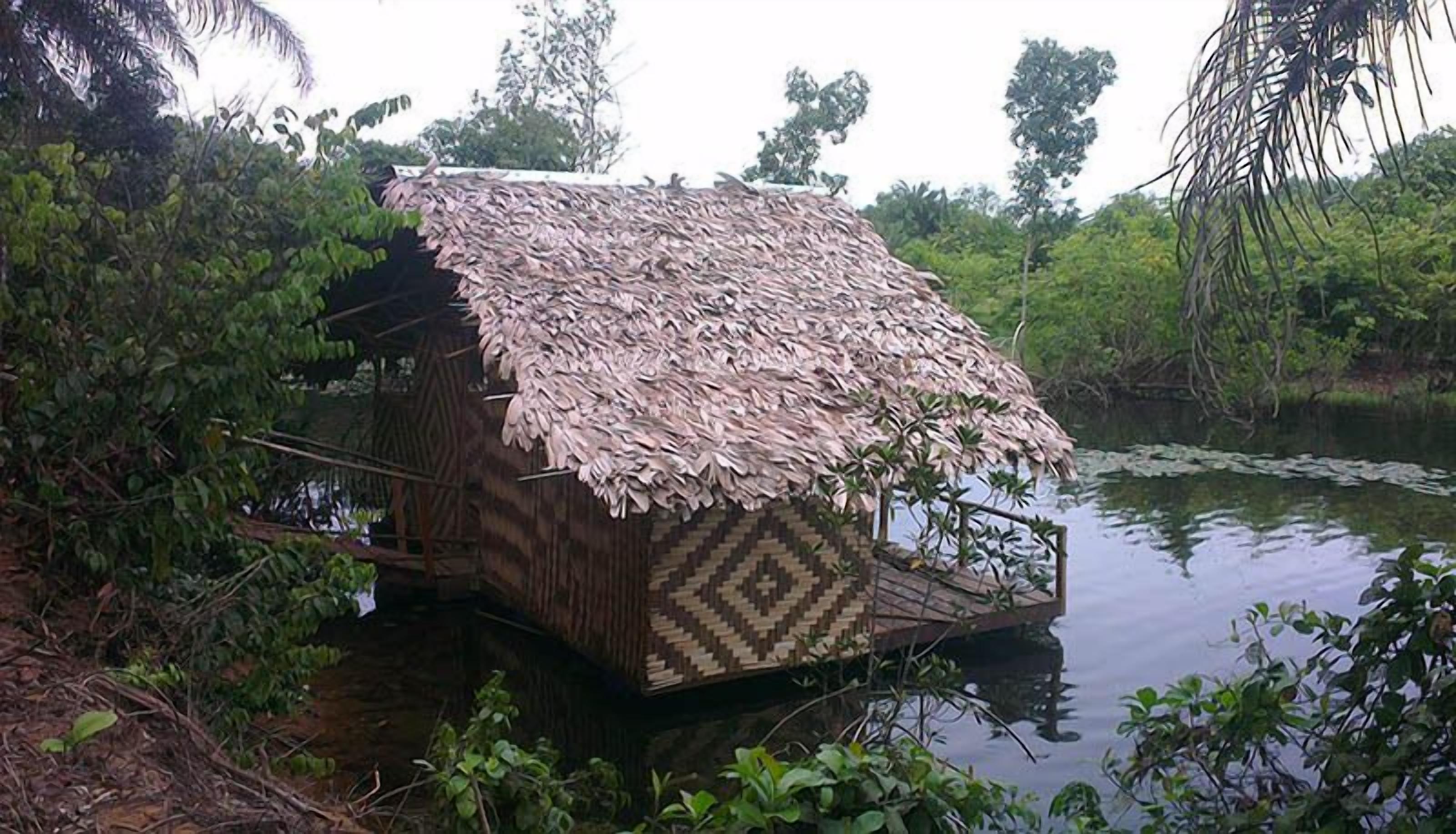 Una cabana amb teulada de palla i paret amb un disseny de línies irregulars, situada sobre una plataforma flotant en un estany rodejat de vegetació densa.
