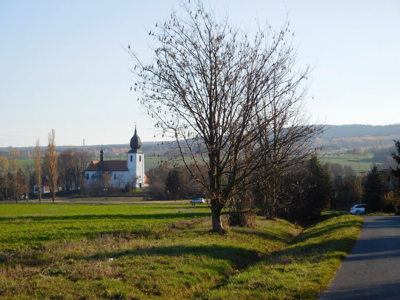 Un paysage rural avec une église blanche surmontée d’un clocher noir, entourée de champs et sous un ciel dégagé.