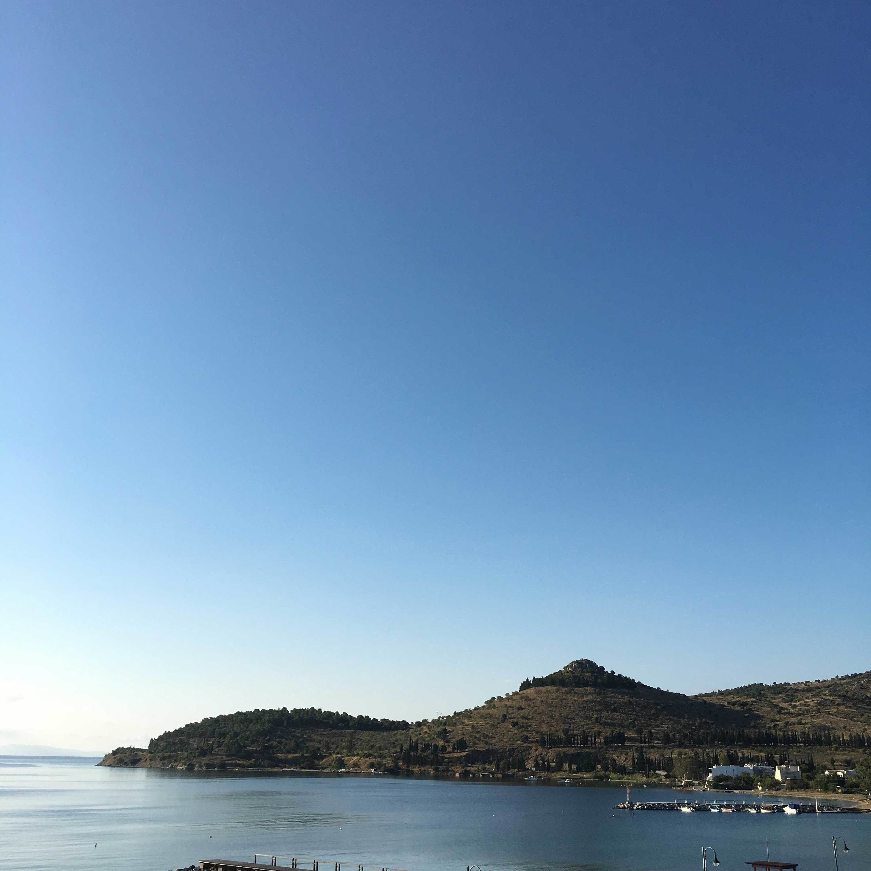 Eine Küstenlandschaft mit blauem Himmel, hügeligem Gelände und einem Gewässer mit Booten.