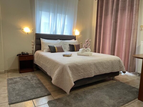 A neatly made bed with a white bedspread, a bedside table with a lamp, and a window with sheer curtains. - Hotel Karagianni (Volos)
