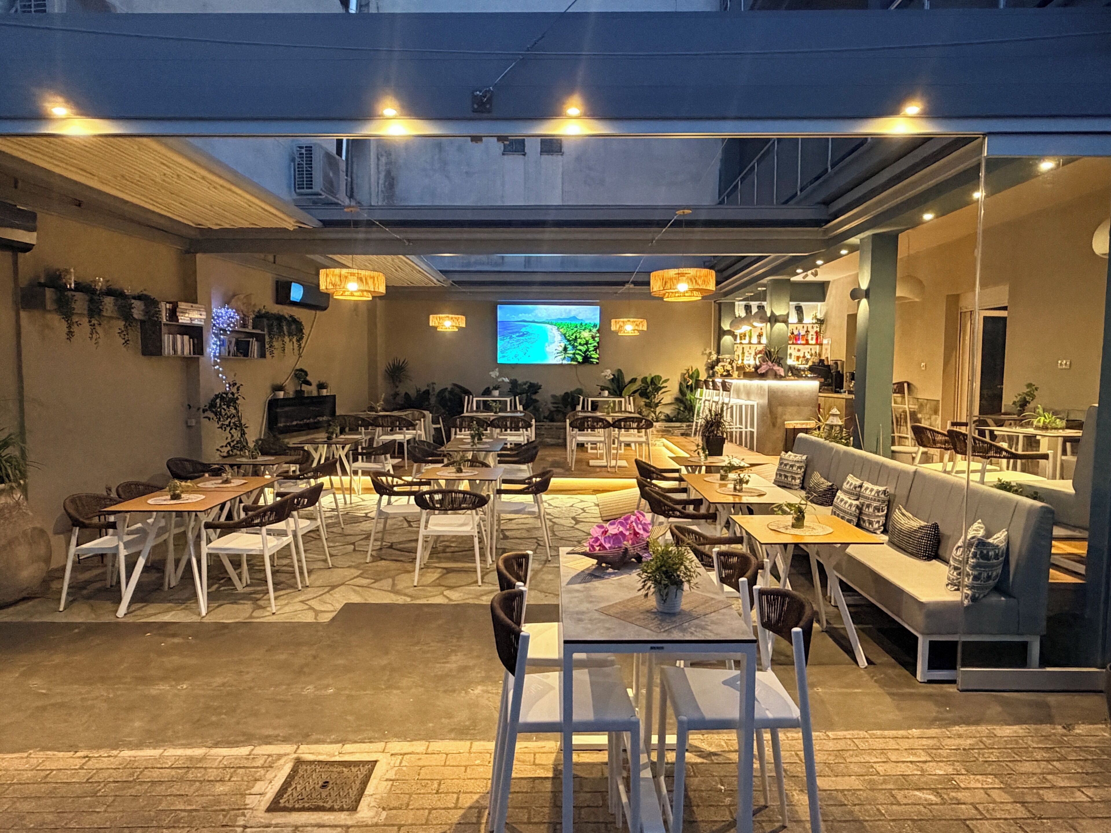 An outdoor dining area with multiple tables and chairs, a bar area, and a television screen displaying a landscape.