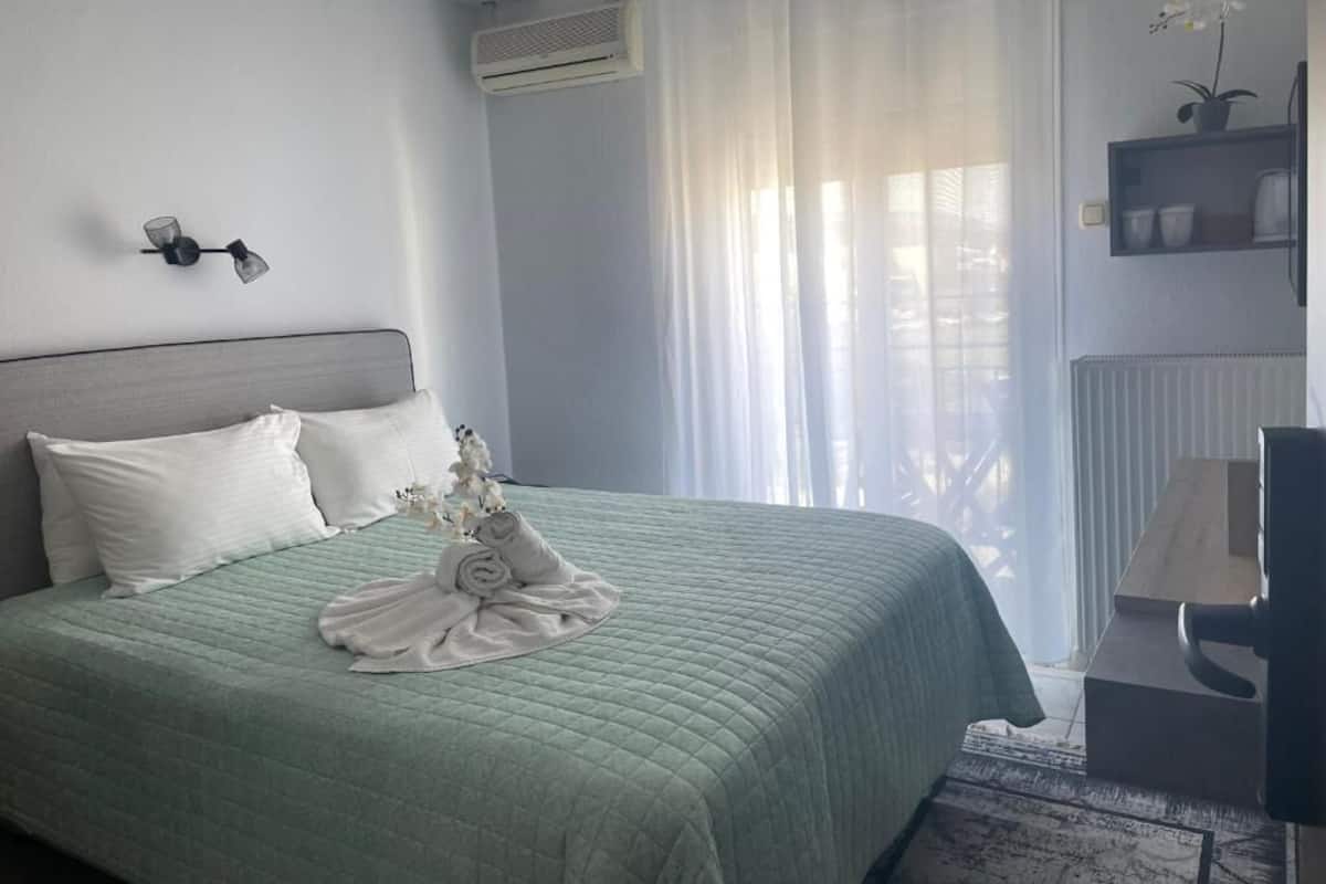 A neatly made bed with a green bedspread and white pillows, a small table with a lamp, and a potted orchid on the windowsill.