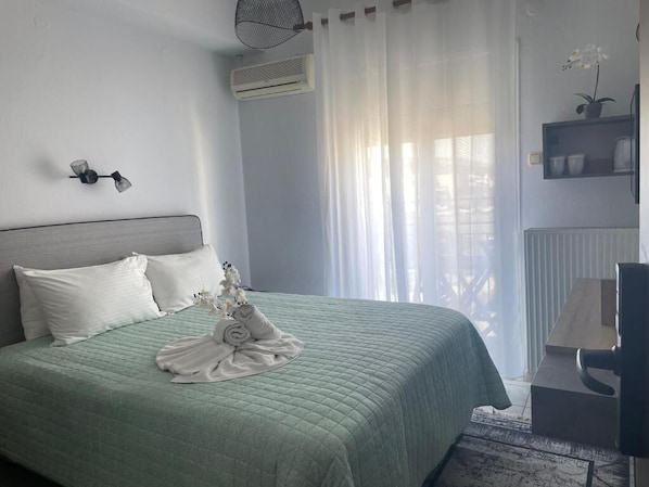 A neatly made bed with a green bedspread and white pillows, a small table with a lamp, and a potted orchid on the windowsill. - Hotel Karagianni (Volos)