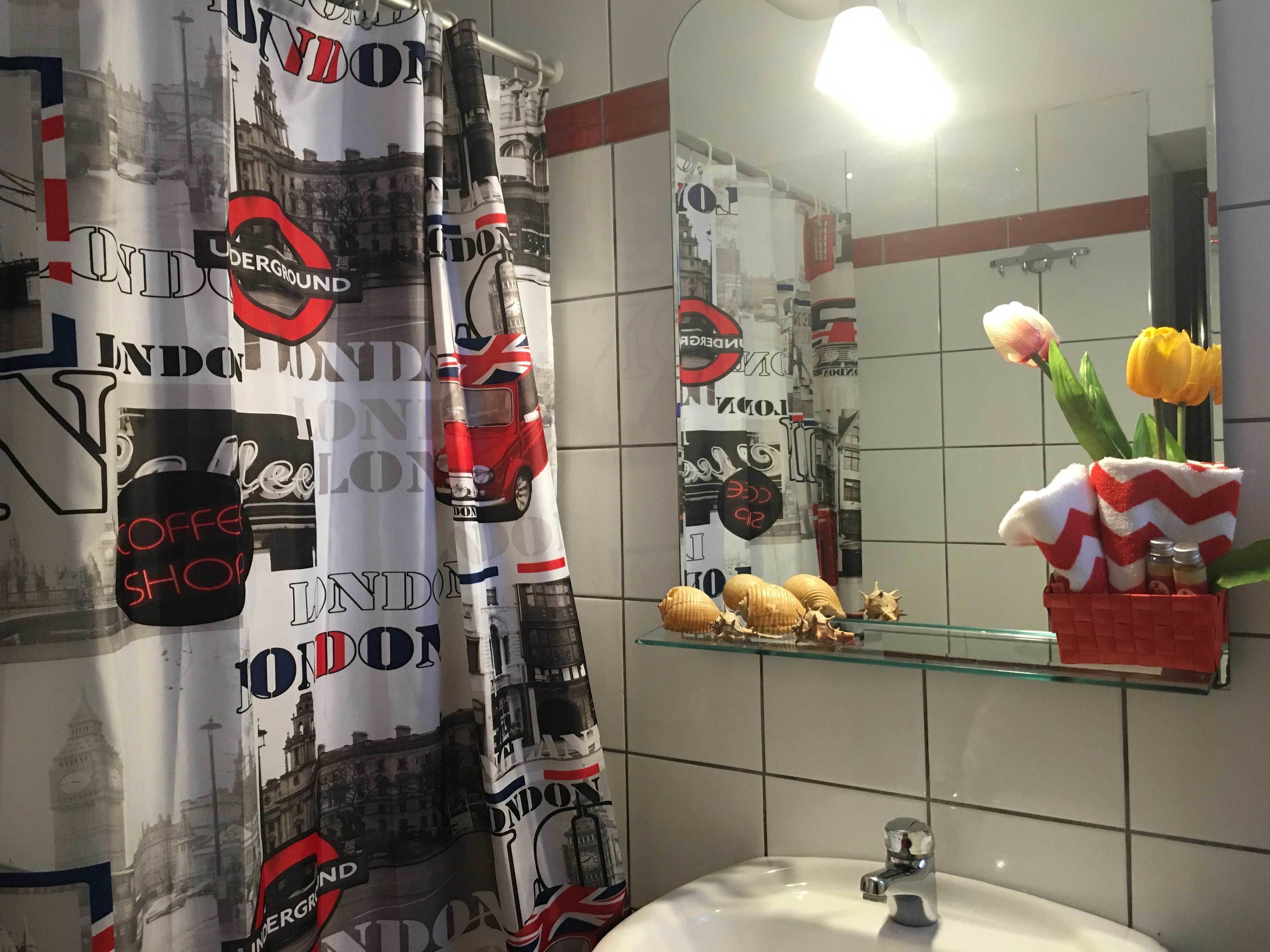 Un baño con cortina de ducha temática de Londres, un lavabo, un espejo y una repisa de cristal con un jarrón de tulipanes.