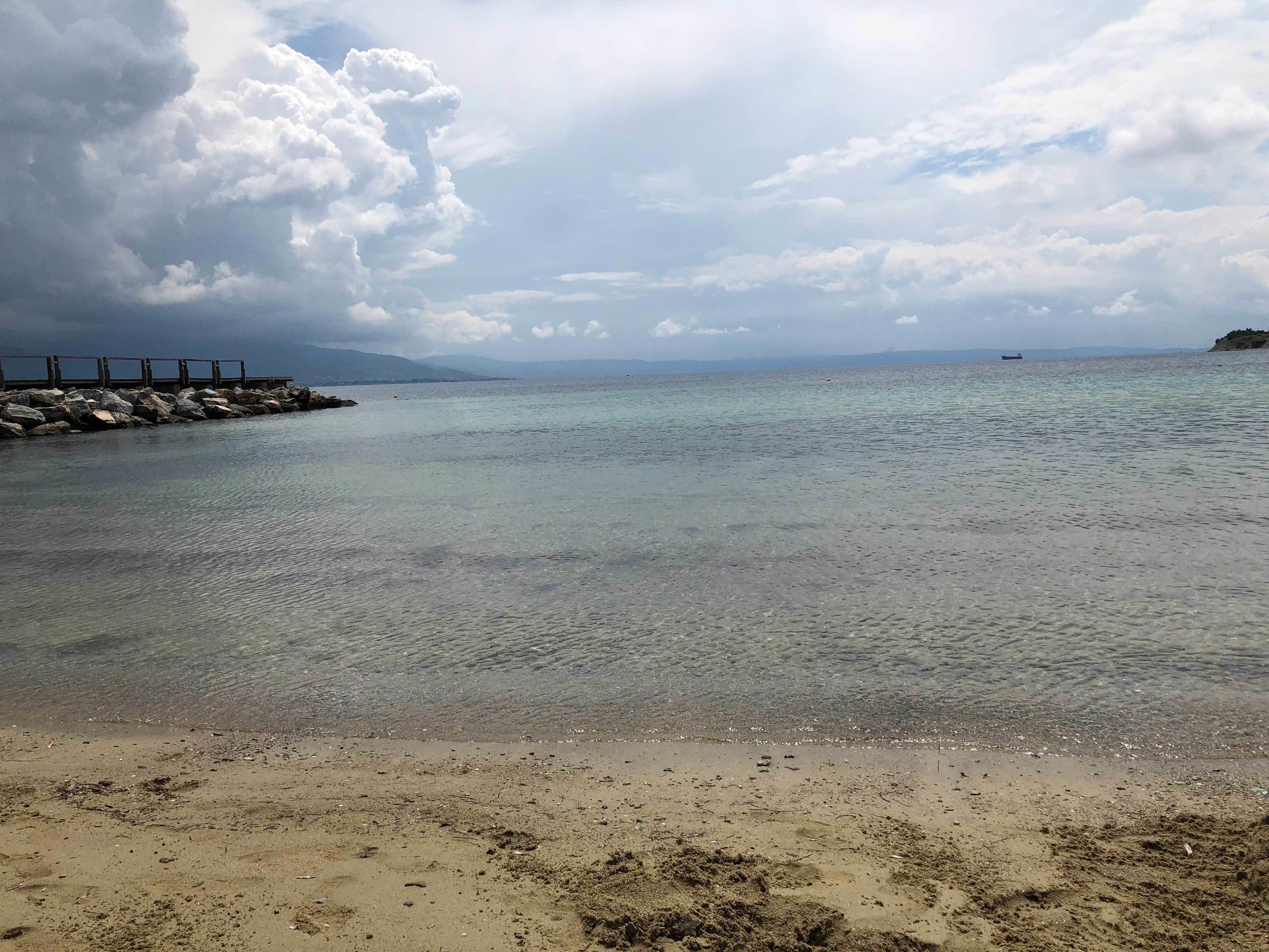 Una playa con agua cristalina, un muelle de madera y montañas a lo lejos.