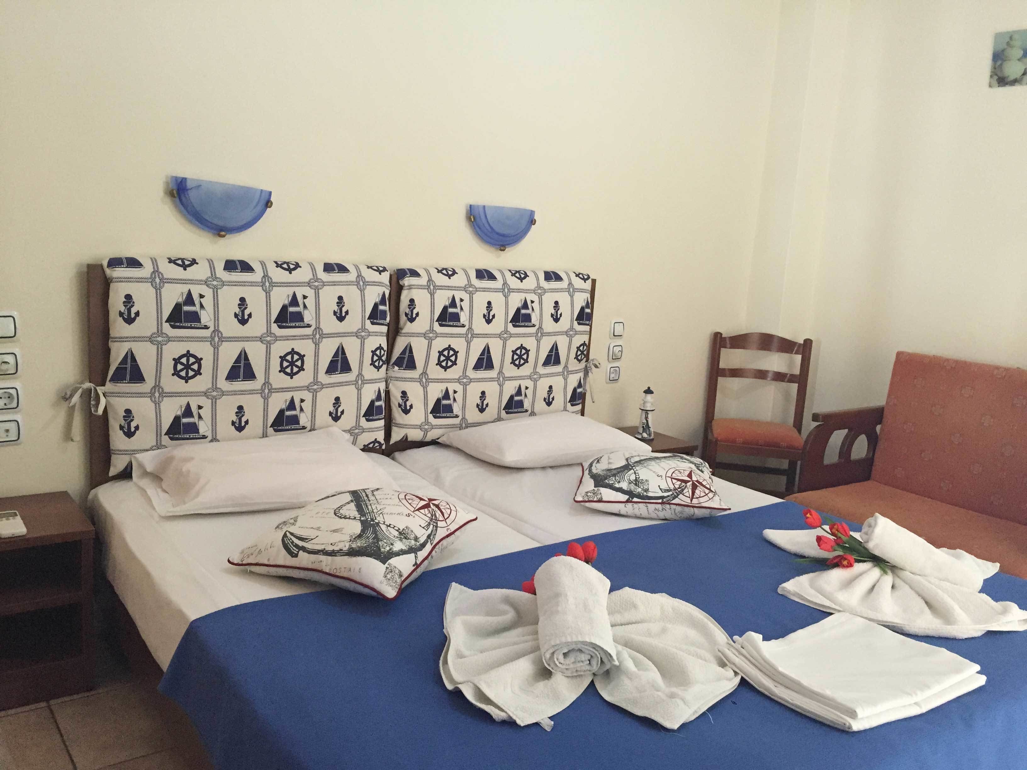 Habitación de hotel con cama y cabecera y almohadas con motivos náuticos, colcha azul y ropa de cama bien tendida, con toallas y un ramo de flores.
