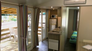A compact kitchen with a microwave, refrigerator, and sink. A small bed with a colorful blanket is visible in the adjacent room. - Camping Lei Suves- Greenchalets (Roquebrune-sur-Argens)
