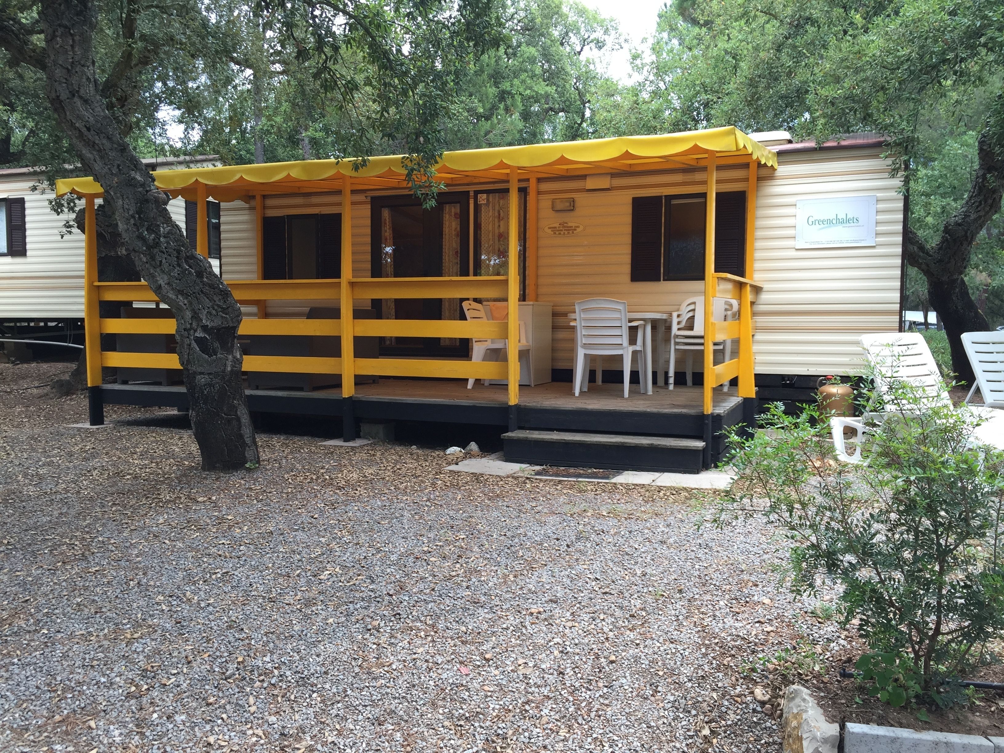 A yellow mobile home with a porch, white chairs, and a sign reading "Greenhalets".