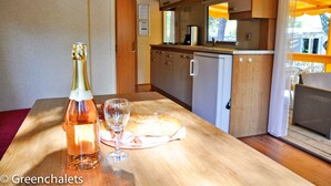 A compact kitchen with wooden cabinets, a small refrigerator, and a dining table with a bottle of wine and a glass. - Camping Lei Suves- Greenchalets (Roquebrune-sur-Argens)