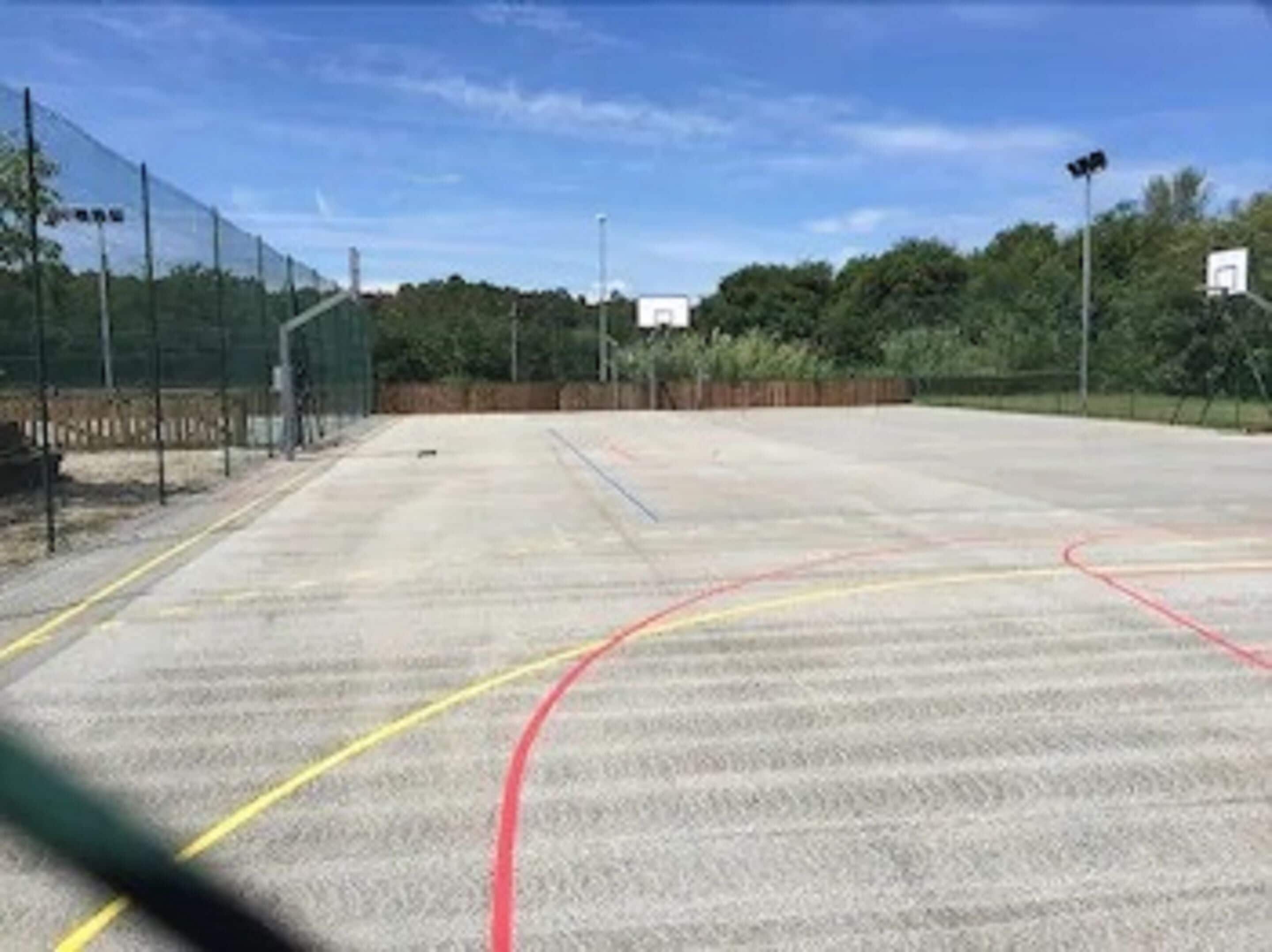 An outdoor basketball court with a fence, trees, and a clear sky.