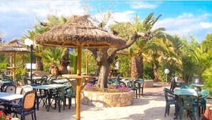 An outdoor dining area with thatched umbrellas, palm trees, and outdoor seating. - Camping Lei Suves- Greenchalets (Roquebrune-sur-Argens)