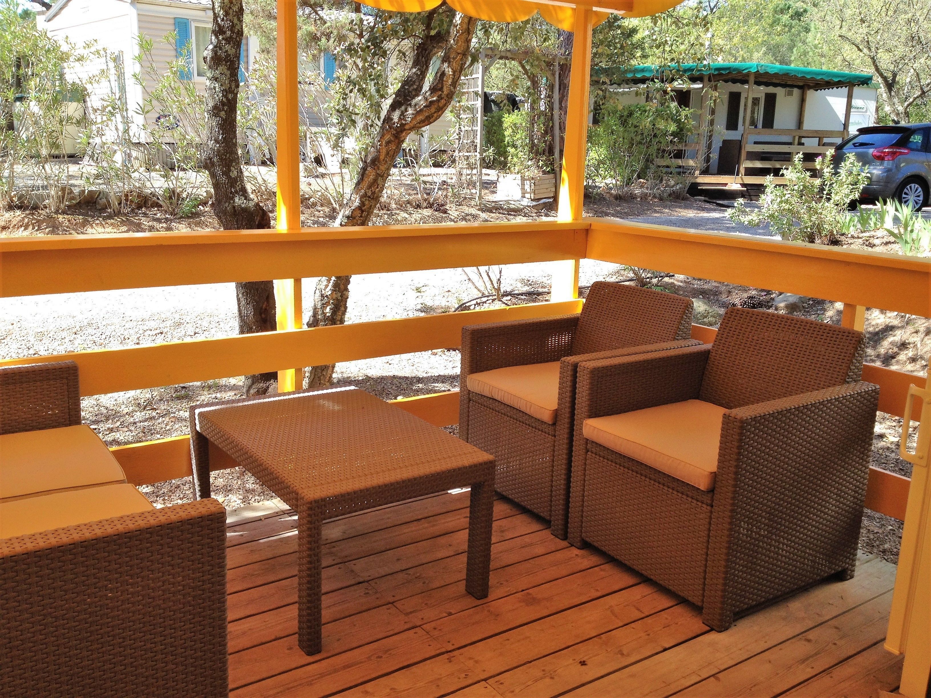 Una terraza de madera con mobiliario de mimbre, incluyendo una mesa y sillas, con vistas a un estacionamiento con un vehículo y árboles.