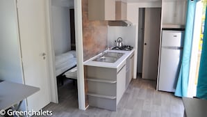 A compact kitchen with a sink, stove, and refrigerator, adjacent to a bedroom with a bed and a door leading to another room. - Camping Lei Suves- Greenchalets (Roquebrune-sur-Argens)