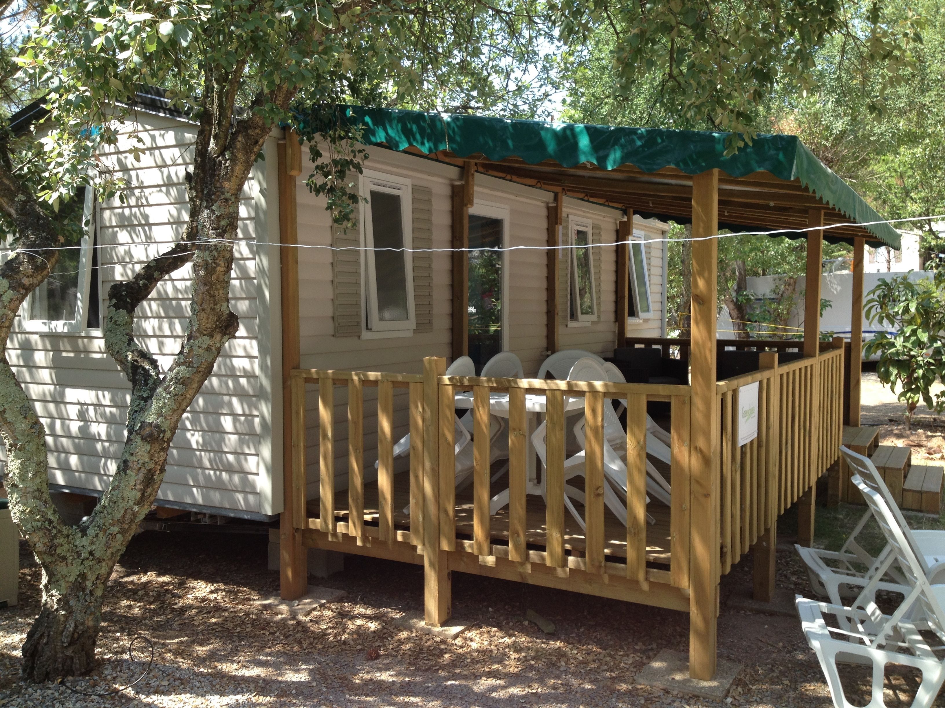 Eine Holzterrasse mit einem grünen Markisenvorzelt, weißen Kunststoffstühlen und einem kleinen Tisch.
