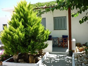 Un pequeño jardín con un árbol de coníferas, un patio con mesa y sillas, y un edificio blanco con contraventanas.