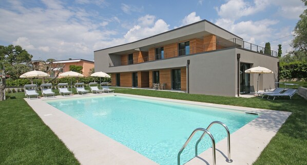 Una casa moderna de dos pisos con piscina, sillas de descanso y sombrillas.