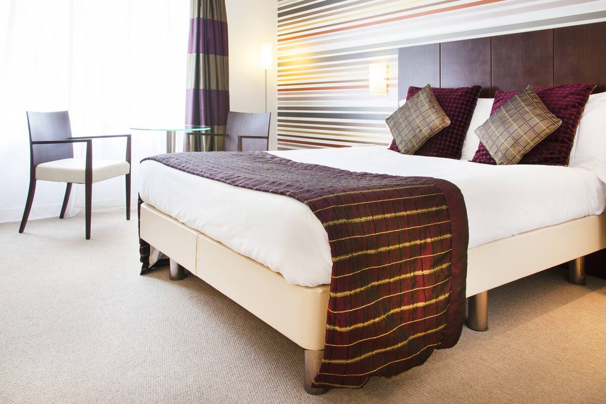 A hotel room with a bed, a chair, a glass table, and a wall with horizontal lines.