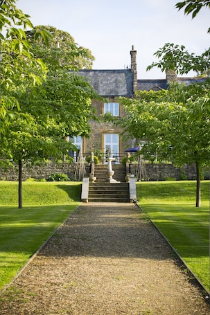 A stone building with a central staircase leading to a door, surrounded by well-maintained lawns and trees.