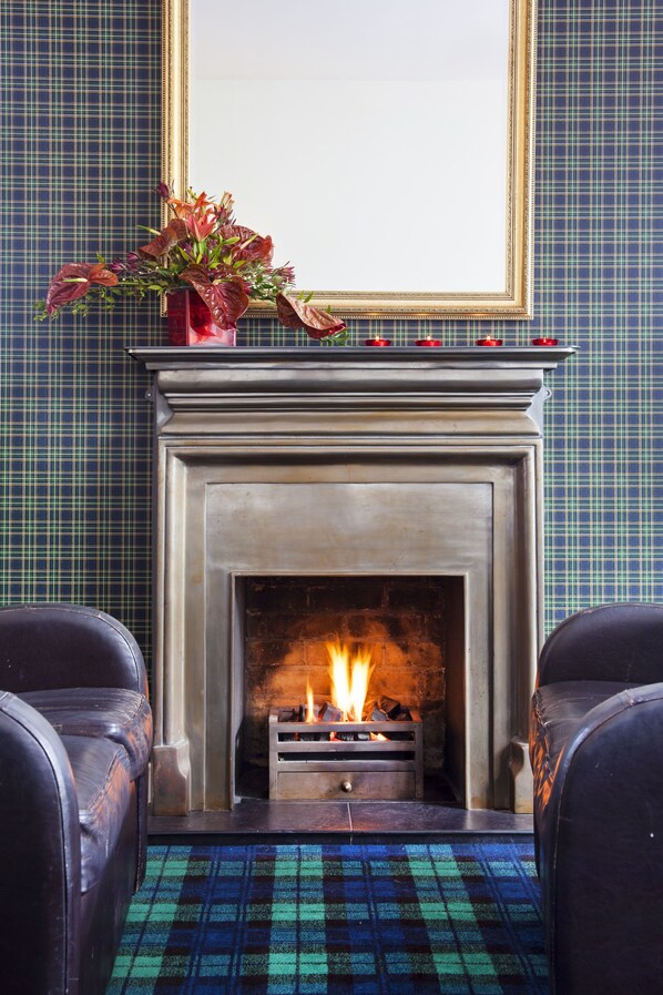 A fireplace with a fire, flanked by two leather chairs, against a wall with a plaid pattern. - Lanes Hotel (Yeovil)