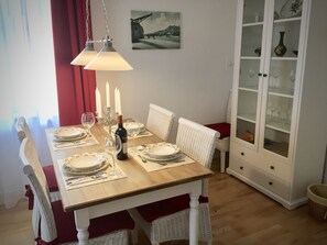 A dining room with a wooden table set for six, white wicker chairs, a cabinet with glass doors, and a painting on the wall.