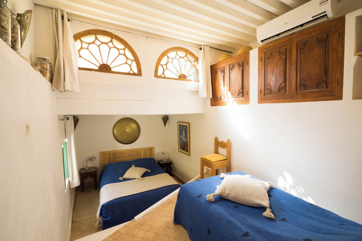 A room with two beds, a wooden cabinet, a wall-mounted air conditioner, and a decorative window.